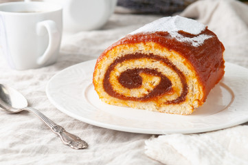 A piece of homemade sponge cake with apple jam. On a white plate. In the background is a white teapot and a mug. Close-up.