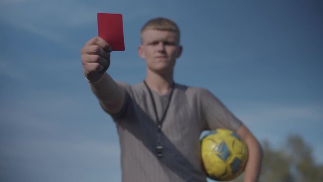 Strict football referee with soccer ball showing red card to send off offending player during game over blue sky background. Soccer referee showing red card indicating dismissal for match to player.