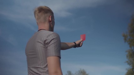 Rear view of determined football referee showing red card and sending-off player after harsh foul committed during match. Soccer referee presenting red card indicating dismissal for match to player
