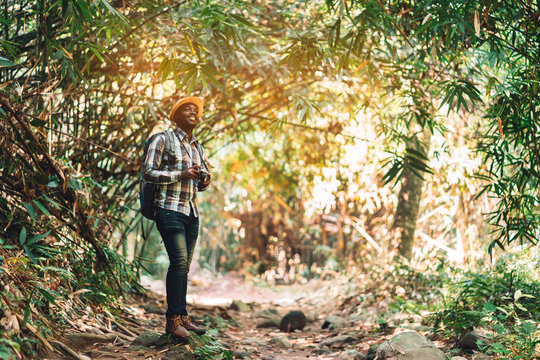 African man traveller holding camera with backpack standing in the green natural forest.