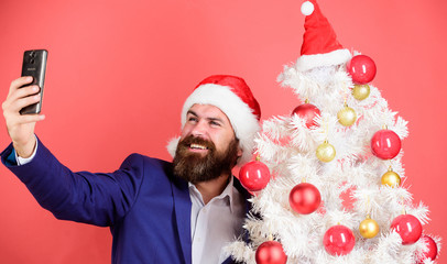 Winter holidays concept. Santa claus takes selfie near christmas tree on red background. Santa holds cell phone. Capturing christmas memories. Man with beard and happy face send christmas greetings