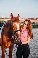 Obraz premium Young smiling woman standing in relax outside with wonderful chestnut horse on bridle and looking away.