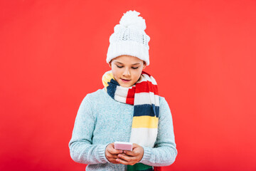 front view of kid in winter outfit using smartphone isolated on red