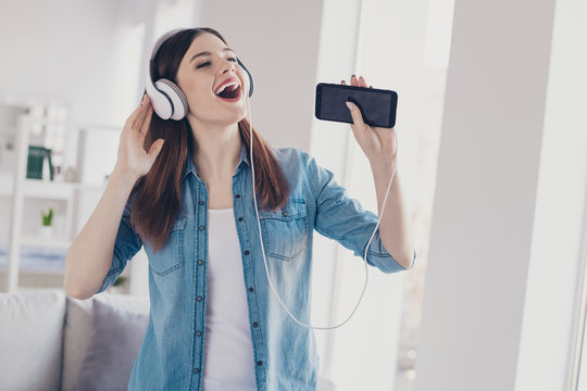 Photo of pretty lady with telephone in hands earflaps on ears singing loudly homey mood standing near sofa wearing jeans clothes apartment indoors