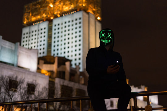 Man wearing scary led green mask looking into the camera with high industrial building in the background