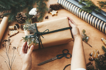 Merry Christmas. Hands holding stylish rustic christmas gift and pine branches, cones, gingerbread cookies,thread, cinnamon, cotton on rural wooden table. Season's greeting