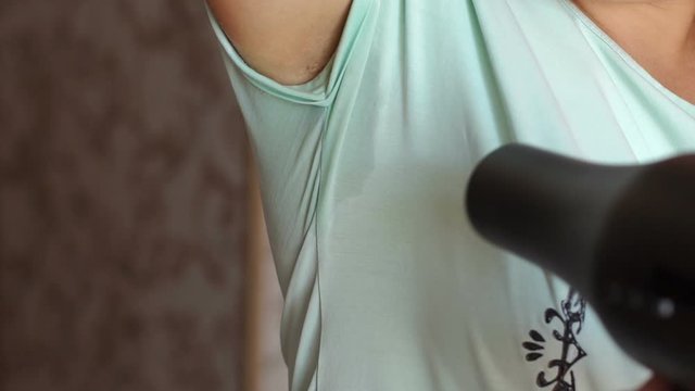 Woman drying wet armpit using hair dryer. Getting rid of sweat and bad smell, hyperhidrosis. Woman with sweat stain on her clothes under armpit is drying her armpit with hair dryer.
