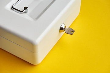 Close up of small white closed cashbox or money box with key in lock on yellow background with copy space  