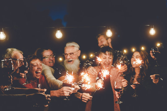 Happy family having at dinner party outdoor - Group of multiracial older and young people celebrating together drinking wine holding fireworks sparklers - Concept of youth and elderly parenthood