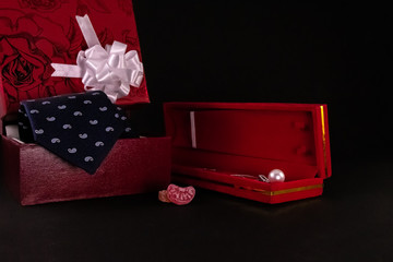 Anniversary gift concept. Beautiful silver pearl necklace in red box, candy wih blue tie and decorative gift box on black background