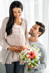 handsome man hugging belly of his pregnant african american woman and holding bouquet