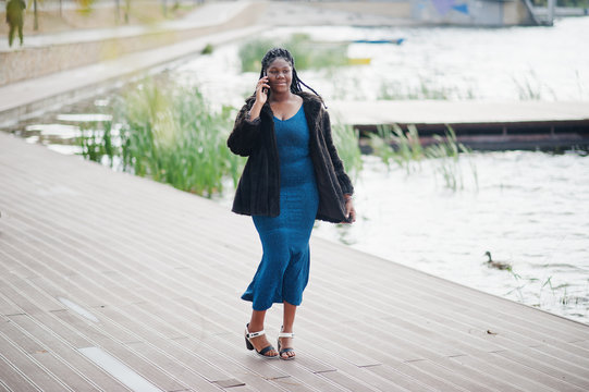 African american dark skinned plus size model posed in a blue shiny dress and black fur coat against lake and speaking by mobile phone.