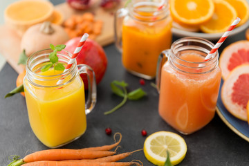 Fototapeta na wymiar food , healthy eating and vegetarian concept - mason jar glasses of orange and carrot juices with paper straws, fruits and vegetables on slate table