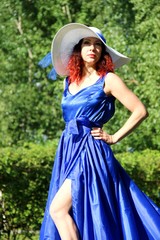 a lady in a blue dress and a gorgeous snow-white hat on a green lawn in the Park. a woman in a hat and a long dress with a train on the skirt on the background of green grass and trees in the city