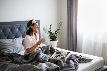 good morning concept - portrait of happy young beautiful woman in pajamas sitting on bed and drinking coffee - obrazy, fototapety, plakaty