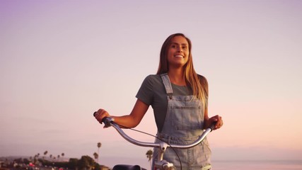 Cute smiling girl walking bike on beach boardwalk at sunset in summertime