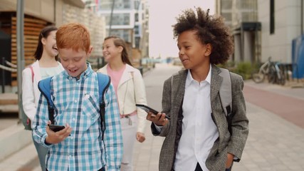 Medium shot of five multiethnic school kids with backpacks going out of school and chatting. Two boys using telephones on foreground