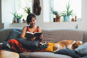 woman sitting sofa and reading book with her dog at home