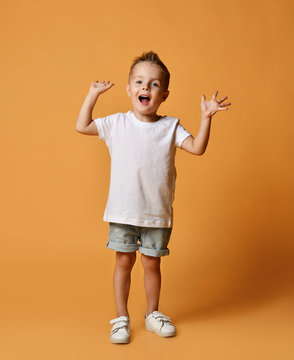 Happy screaming boy kid in white t-shirt, blue jeans shorts and white sneakers yelling with his hands up on yellow