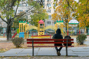 Fototapeta na wymiar young man in dark clothes and a hood sits alone on a bench near the playground