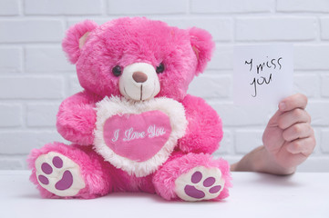 A pink toy bear is sitting on the table and the guy is holding a note in his hand against a white loft. Toy bear on the table close-up. Holiday Greeting Card. Holiday background.
