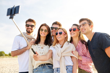 friendship, leisure and people concept - group of happy friends taking picture by smartphone on selfie stick on beach in summer