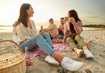 friendship, leisure and fast food concept - group of happy friends eating sandwiches or burgers at picnic on beach in summer