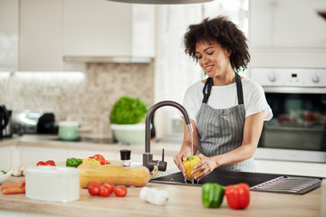 Charming mixed race woman in apron standing in kitchen ad washing yellow paprika in sink. On...