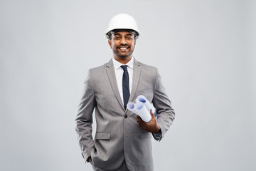 architecture, construction business and people concept - smiling indian male architect in helmet with blueprints over grey background
