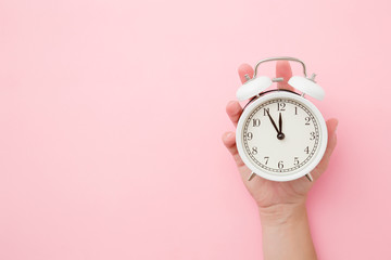 Woman hand holding white alarm clock on light pastel pink background. Time concept. Closeup. Empty...
