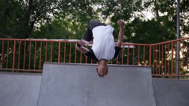 Blonde young guy in in wide black pants and a red bandage on his hair makes a beautiful somersault running on a skate ramp in the park. Footage from the back