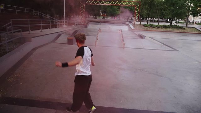 Front footage from high angle view of young guy performing somersaulting jump on sunny summer day while holding red coloured burning smoke bomb on the skate ramp. Slow motion