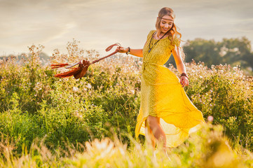 Smiling bohemian girl in yellow dress with guitar on the field at sunset warm light