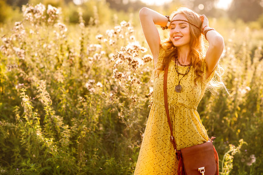 Smiling bohemian girl in yellow dress with guitar on the field at sunset warm light