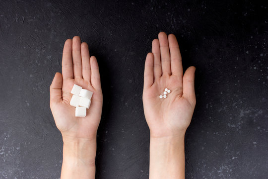  Female hands with sugar and pills on a gray-black background. The choice of sweetener in tablets or regular sugar. An alternative to sugar for diabetics.