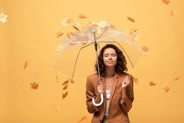 happy curly woman in brown jacket with closed eyes holding umbrella in falling golden maple leaves...