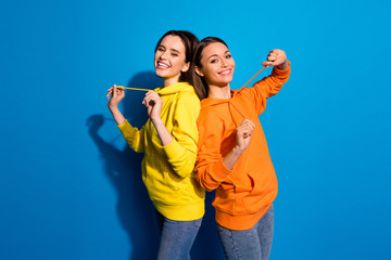 Profile photo of pretty two girlfriends lady standing back-to-back in best good mood wearing casual bright hoodies and jeans isolated vibrant blue color background