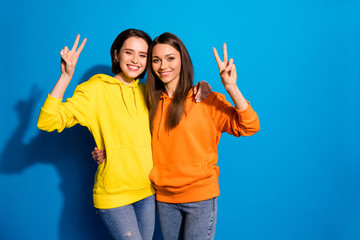 Photo of pretty two girlfriends lady showing v-sign symbols hugging wearing casual bright hoodies...