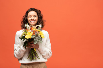 happy woman in autumnal outfit holding bouquet of flowers isolated on orange