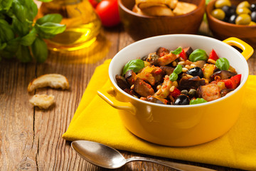 Traditional Italian caponata, served with croutons in a yellow, sunny pot. Decorated with basil and pine nuts.