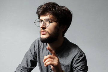 Portrait of young bearded guy with disheveled hair and glasses, looking away and talking, pointing up with finger on background of grey.