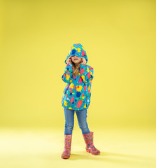 A full length portrait of a bright fashionable girl in a raincoat holding an umbrella of rainbow colors on yellow studio background. Autumn and spring fashion for kids. Cute stylish blonde girl.