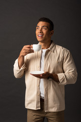smiling mixed race man in beige shirt looking away and holding cup of coffee and saucer on black background