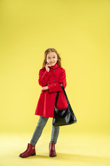 A full length portrait of a bright fashionable caucasian girl in a red raincoat holding a black bag on yellow studio background. Autumn and spring fashion for kids. Cute stylish blonde girl.