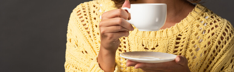panoramic shot of woman in yellow knitted sweater holding cup of coffee and saucer on black background