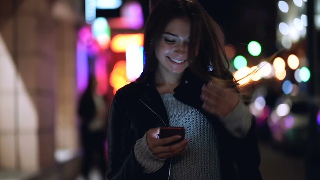 Attractive young woman walking at the night city with smartphone in the hands. She is happy and smiling. Slowmotion. Handheld shot. 