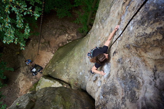 Young female rock climber climbing on cliff with safety harness and rope. Woman overcoming difficulties on rocks. Extreme summer outdoors on difficult climbing route on rock. Sport in nature. Top view
