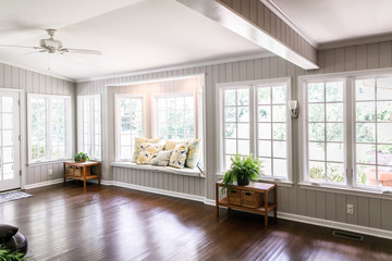 Large and open living room den sun room with windows on two sides and lots of natural light flowing in. There is a window seat on one side and a leather couch and plant on the other. - Powered by Adobe