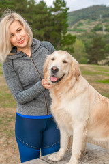 A girl in sportswear caresses a golden retriever dog and looks with a smile. In a coniferous forest against a background of trees and fallen needles
