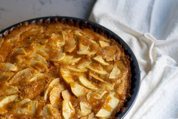 Apple pie in a round shape, decorated with fan-laced apple slices from the top, next to a cup of hot tea, autumn leaves, all on a light, marbled gray background. 